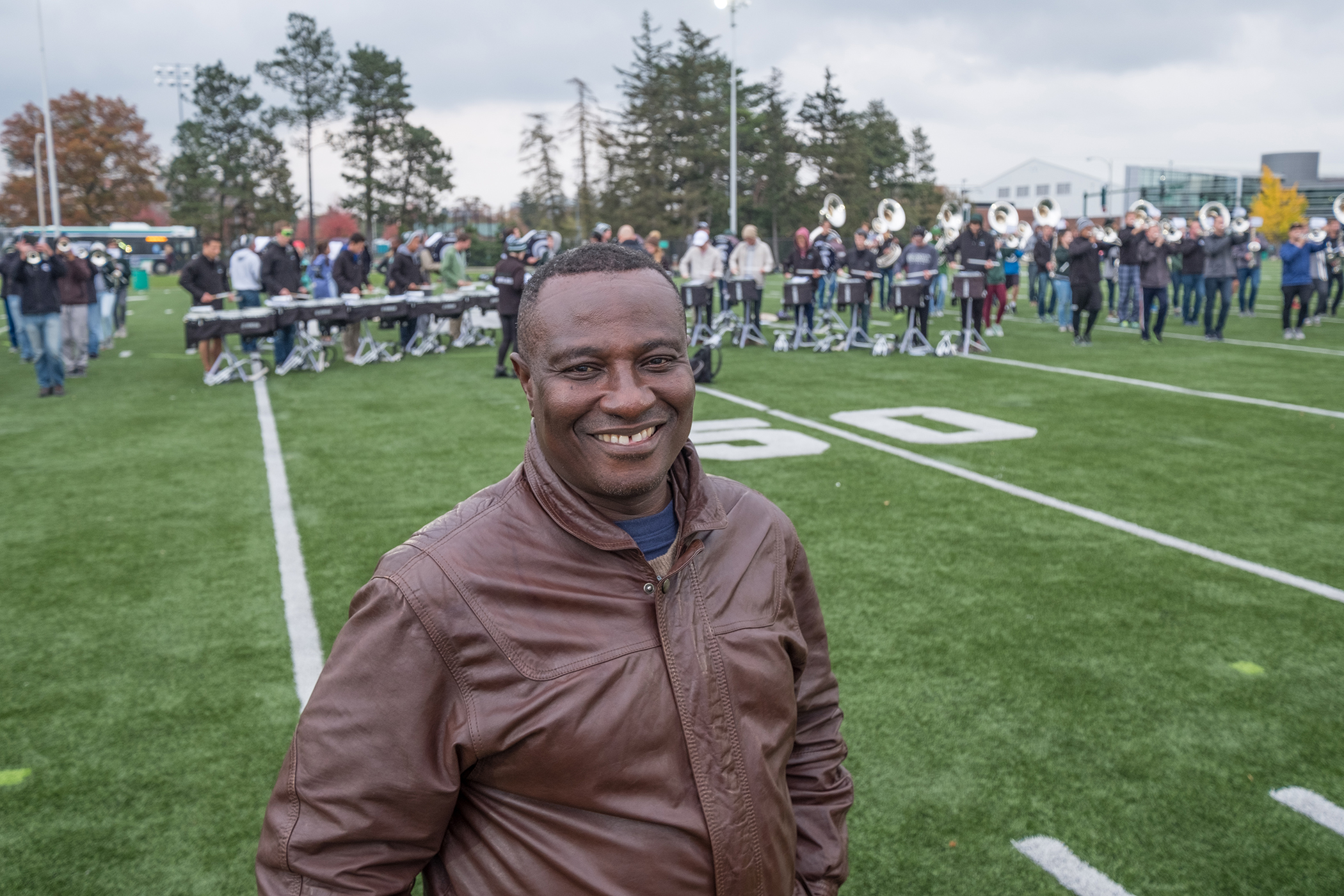 Photo of Ben Ayettey smiling at the camera on a green field, with band members practicing behind him.